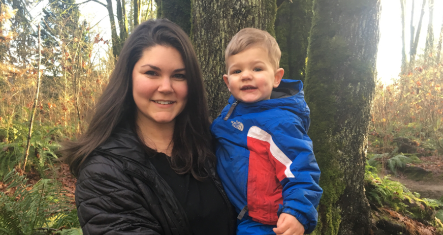 Smiling woman holding a child in a forest with sunlight filtering through the trees.