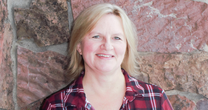 Woman with blonde hair in a red plaid shirt smiling in front of a stone wall.