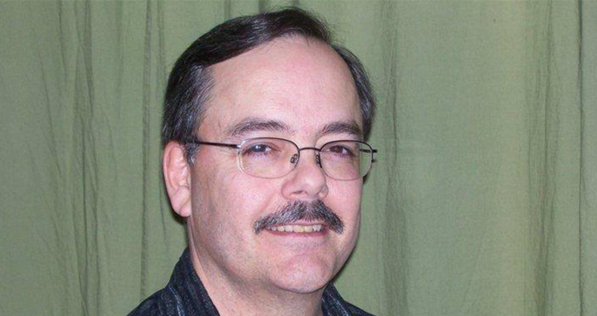 A man with glasses and a mustache smiles in front of a green curtain.