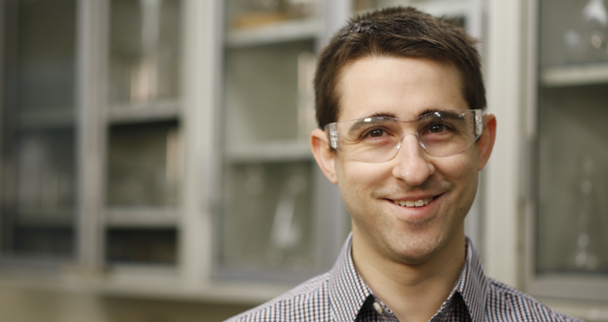 A man wearing safety goggles smiling in a laboratory setting.