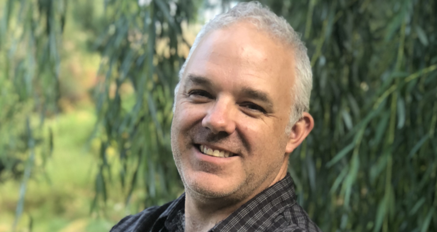 A man with short gray hair smiling outdoors with trees in the background.