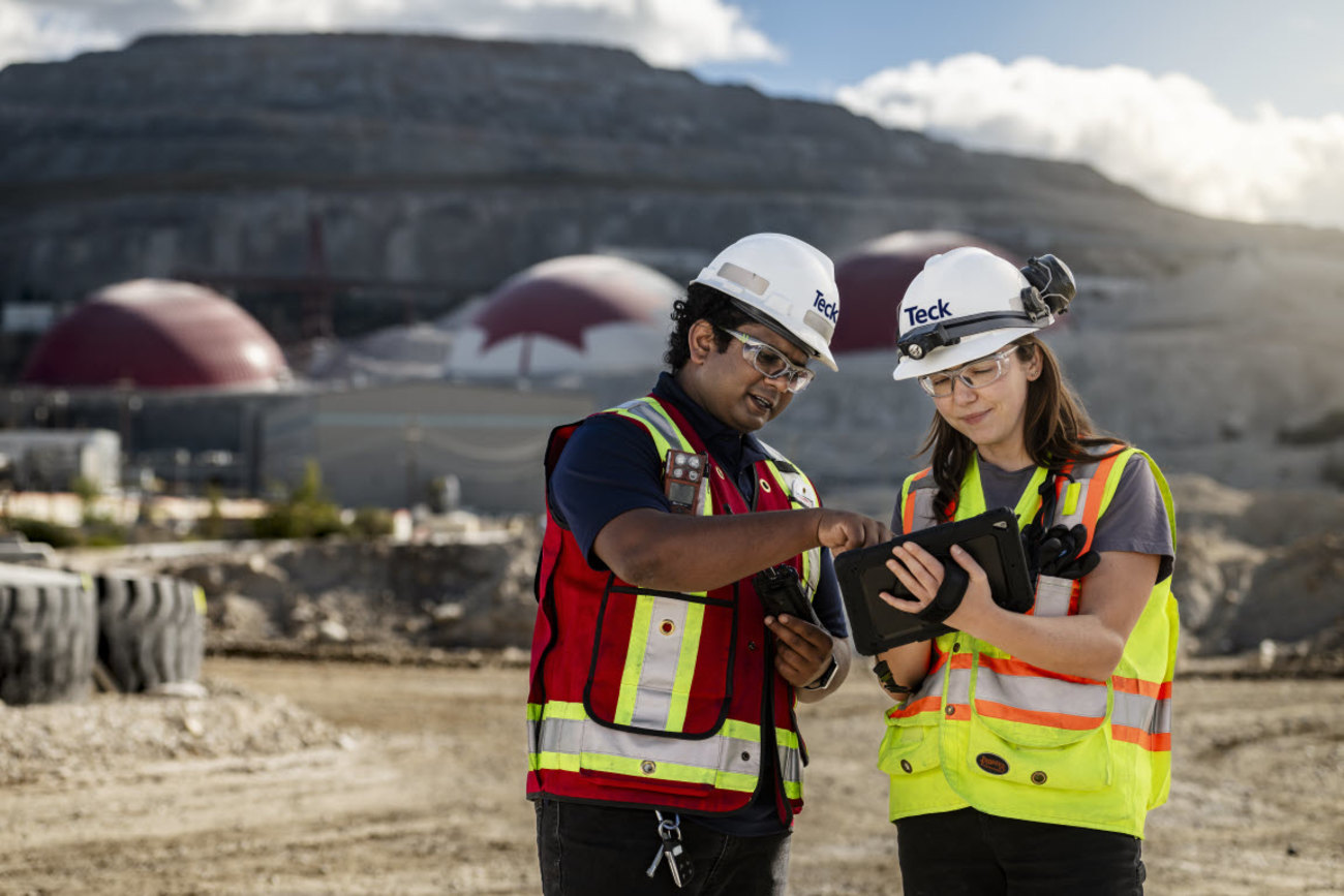 Two engineers in safety gear use a tablet at a mining site.