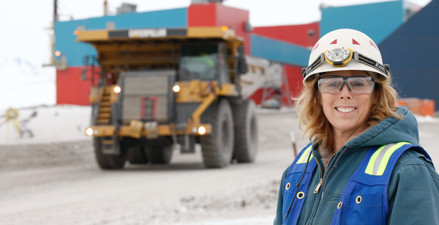 People Make it Possible Worker in safety gear smiles with a mining truck and industrial buildings in the background.