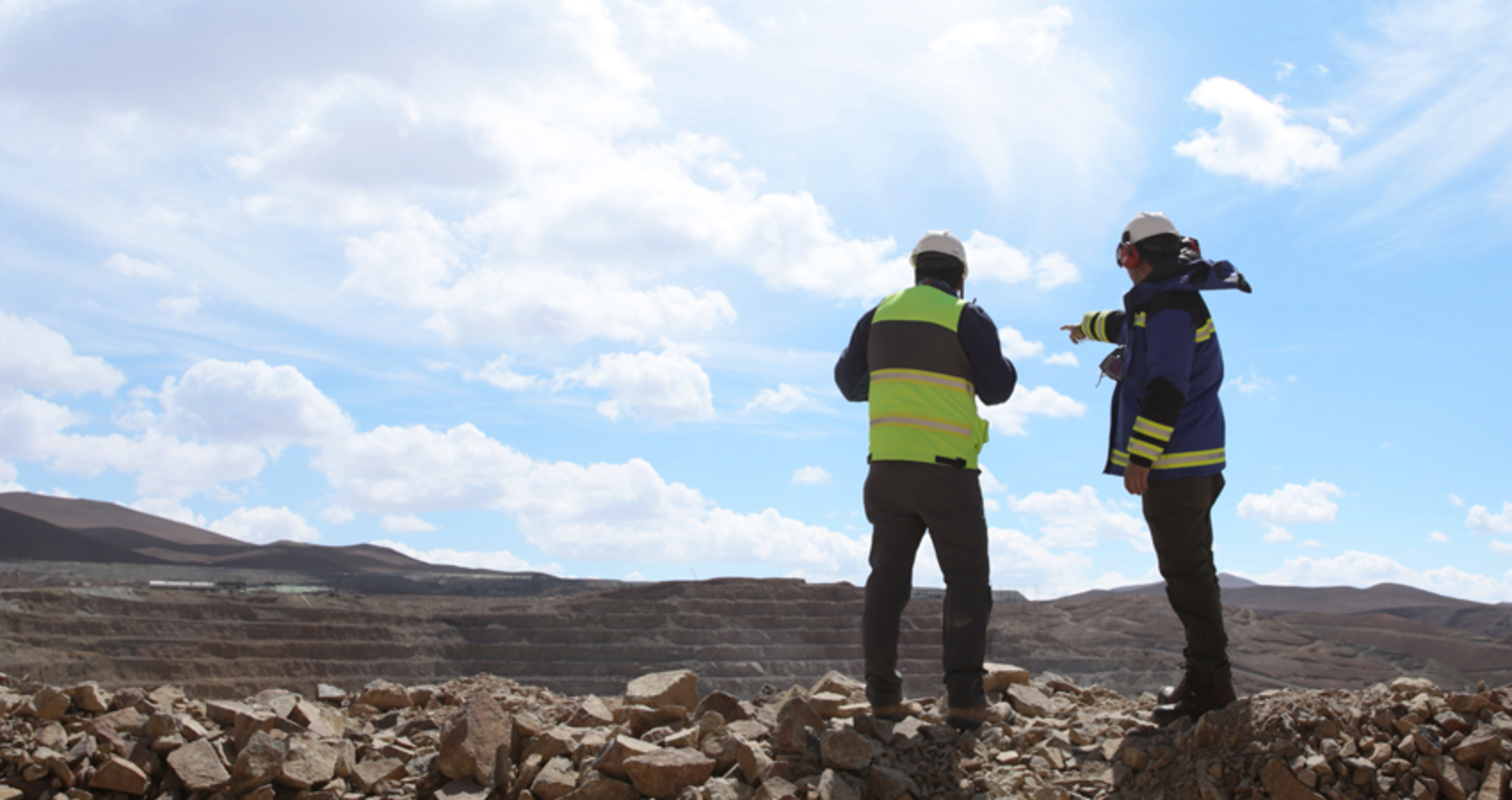Two workers in reflective gear, standing on rocky terrain, pointing and discussing the landscape.