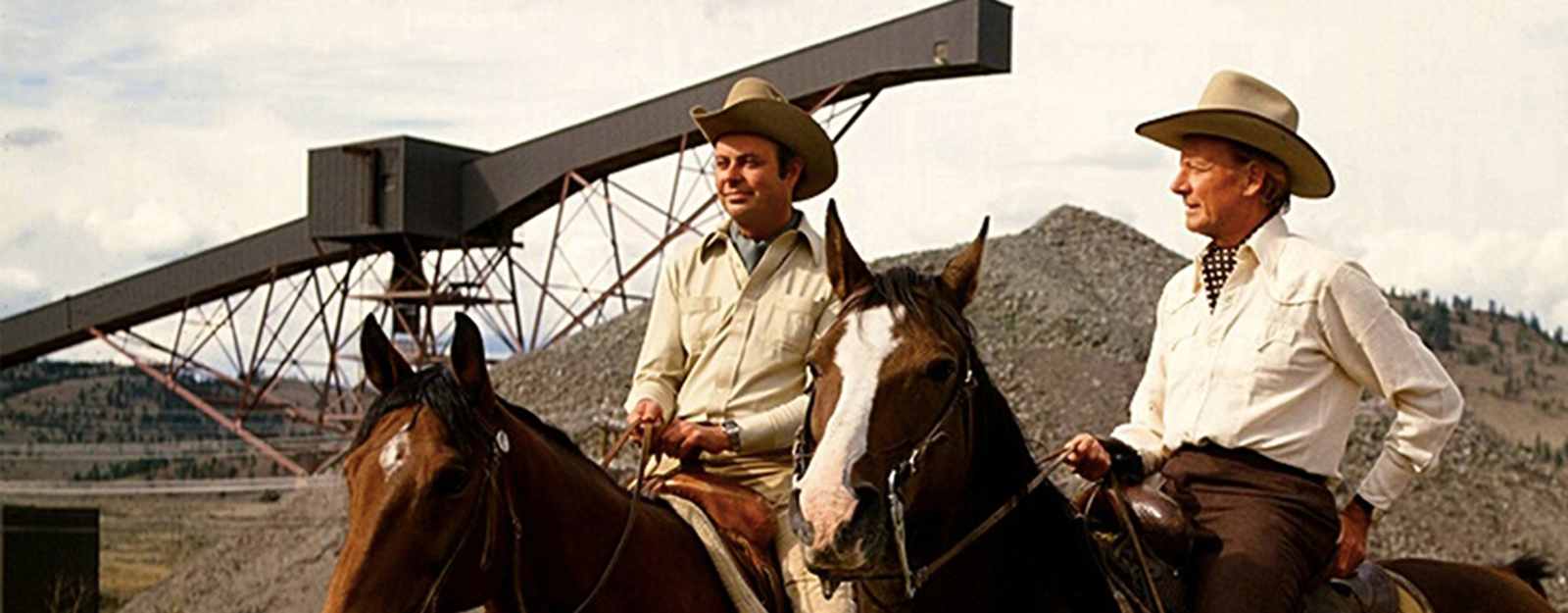Two men ride horses near industrial machinery and rocks, wearing cowboy hats and shirts.