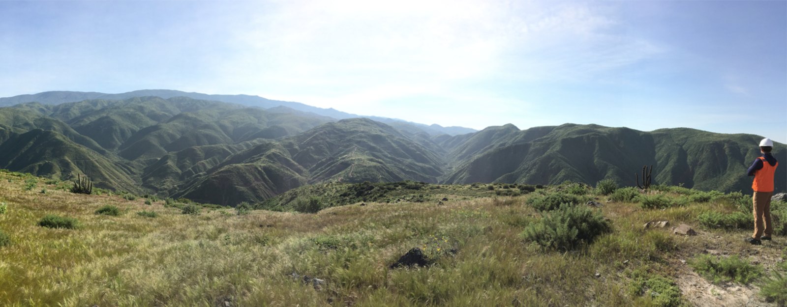 Person in orange vest stands on grassy hill overlooking vast, mountainous landscape.
