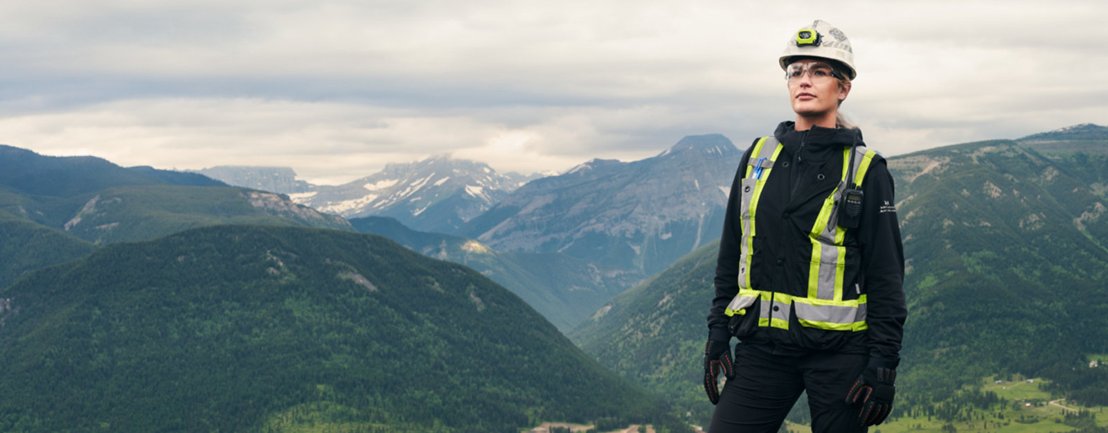 Worker in safety gear and helmet stands with mountains in the background.