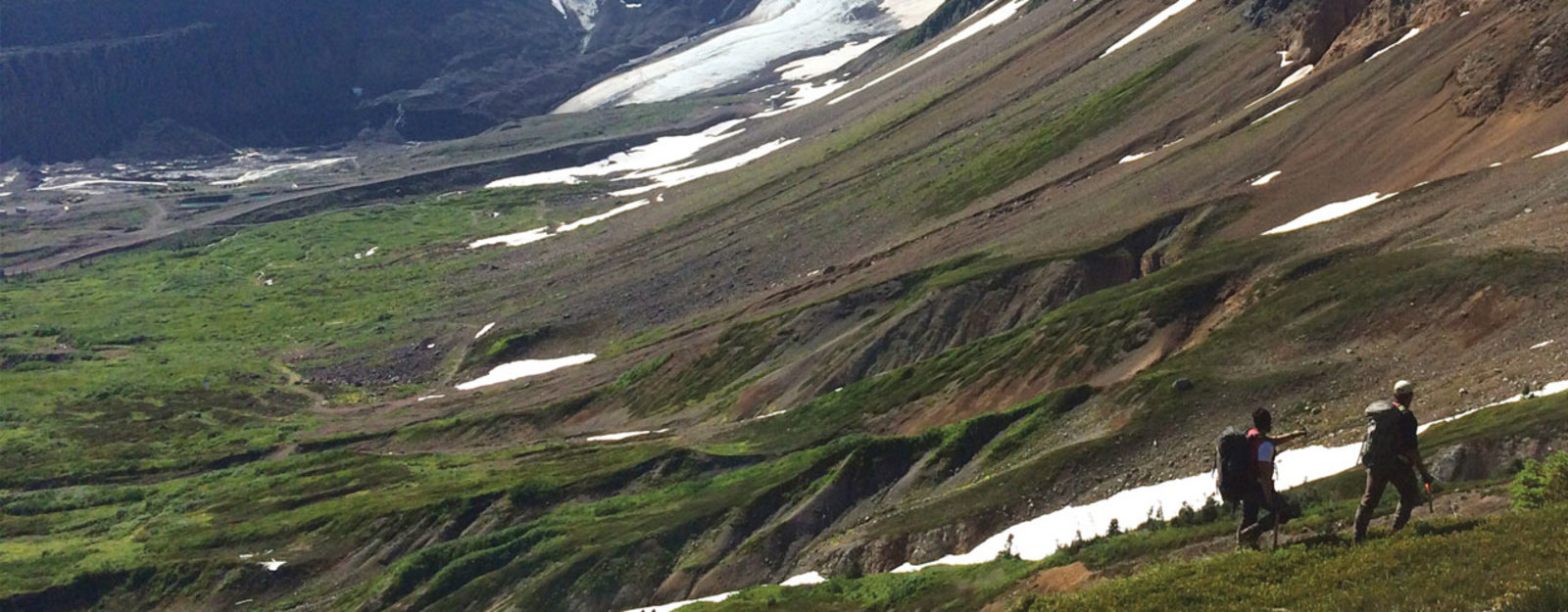 Two hikers in a mountainous area with patches of snow and green vegetation.
