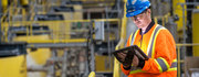 Worker in safety gear and hard hat uses a tablet in an industrial setting.