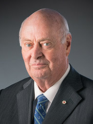 Older man in suit and tie, looking directly at the camera against a gray background.