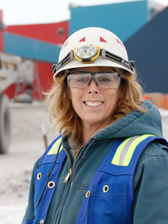 A worker in safety gear and helmet smiles at an industrial site with colorful buildings behind.