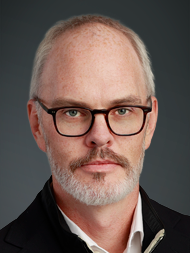 Man with glasses and beard, wearing a dark jacket, against a dark gray background.