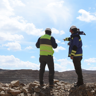 Two people in safety gear at a rocky site, one pointing towards a distant landscape under blue sky.