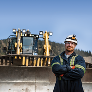 Man in work gear stands in front of large construction equipment with mountains in the background.