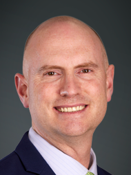 Smiling bald man in suit and tie against a dark background.