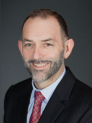 A smiling man in a suit with a red tie, against a dark gray background.