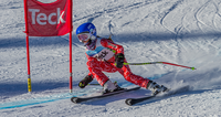 Young skier in red suit and blue helmet racing around a gate on a snowy slope.