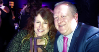 A smiling woman holding an award and sitting next to a smiling man in a suit and tie at an event.
