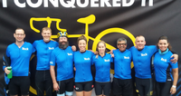 Group of cyclists wearing blue jerseys standing together in front of 'I Conquered It' banner.