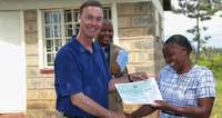 Man in blue shirt handing a certificate to a smiling woman outside a stone building.