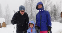 A family of three in winter coats and hats stand in the snow with trees in the background.
