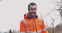 Man in an orange reflective jacket standing outdoors in a forested area, looking at the camera.