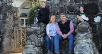 A family of four sits on a stone bench outside a house, smiling at the camera.