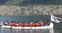 Canoe with a Teck flag carrying a group of paddlers in red life jackets across a scenic lake.
