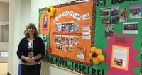 A woman standing beside a colorful bulletin board about healthy foods in a hallway.