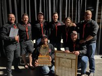 A group of people in black shirts pose with trophies and certificates in front of dark curtains.