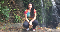 Woman crouching in front of a waterfall with greenery, wearing a colorful shirt and black pants.