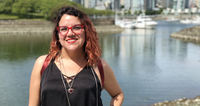 Smiling person with red glasses standing near water with boats and buildings in the background.