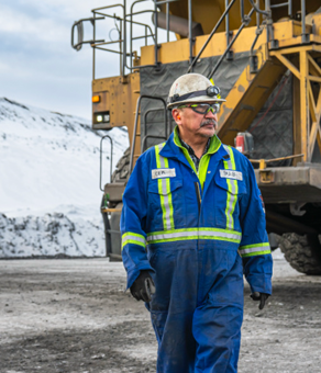 Worker in blue coveralls and helmet stands by large mining truck in snowy landscape.