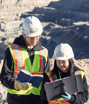 Two construction workers in safety gear reading documents at a mining site.
