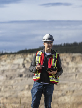 Person in safety gear with tablet, standing outdoors near rocky terrain under cloudy sky.