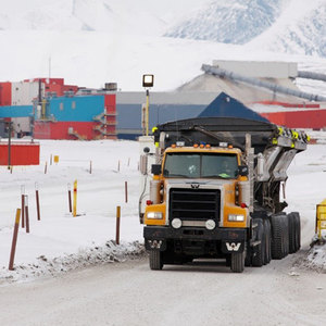 A large yellow truck on a snowy road near colorful industrial buildings and snowy mountains.