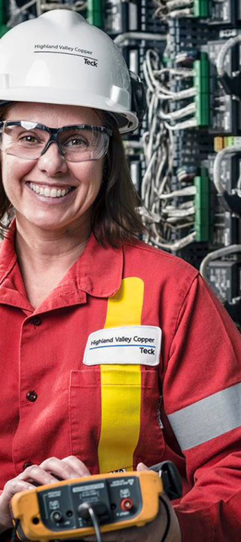 Smiling worker in safety gear holding equipment in front of electronic panels.