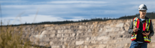 Person in safety gear with a tablet standing in front of a large rock quarry under a blue sky.