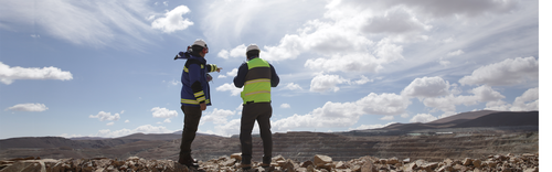 Two workers in safety gear surveying a rocky landscape under a cloudy sky.