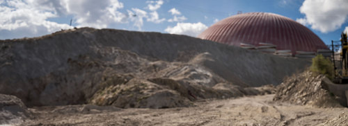 Large red dome structure behind a dirt hill under a cloudy sky.