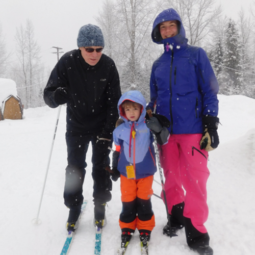 A family of three dressed in winter gear standing together in the snow with skiing equipment.