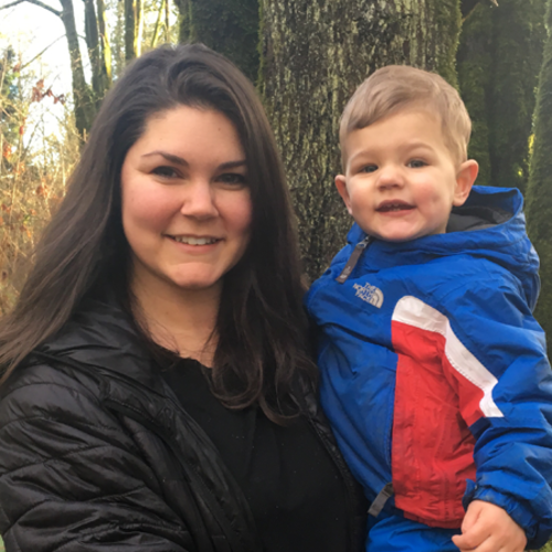 A woman holding a smiling child in a blue jacket, standing in front of trees.
