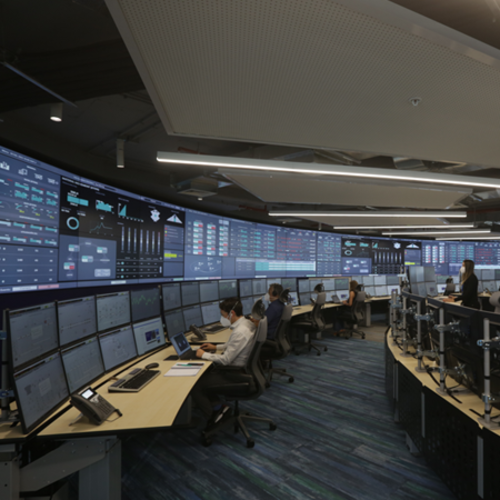 People working at computer workstations in a modern control room with large monitors on the walls.