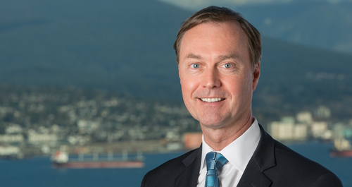 Smiling man in suit and tie, with a city and mountain view in the background.