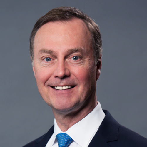 Smiling man in a suit with a blue tie, photographed against a plain gray background.