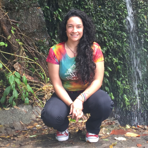 Smiling woman in a tie-dye shirt squatting near a waterfall in a lush, green forest setting.