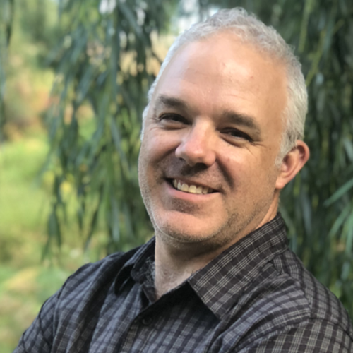 Smiling man with short grey hair and a plaid shirt standing in front of greenery outdoors.