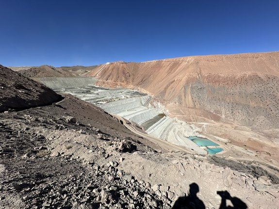 Large open-pit mining area with terraced slopes and small blue pond under a clear blue sky.