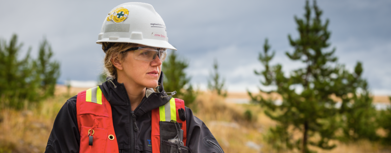 Person in safety gear and helmet, outdoors with trees and cloudy sky in the background.