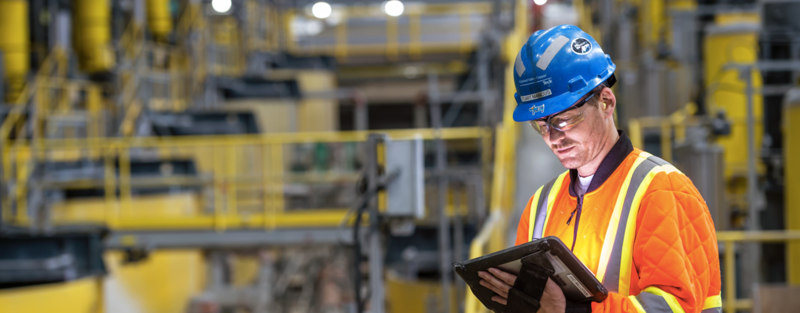 Worker in safety gear and hard hat using a tablet in a large industrial facility.